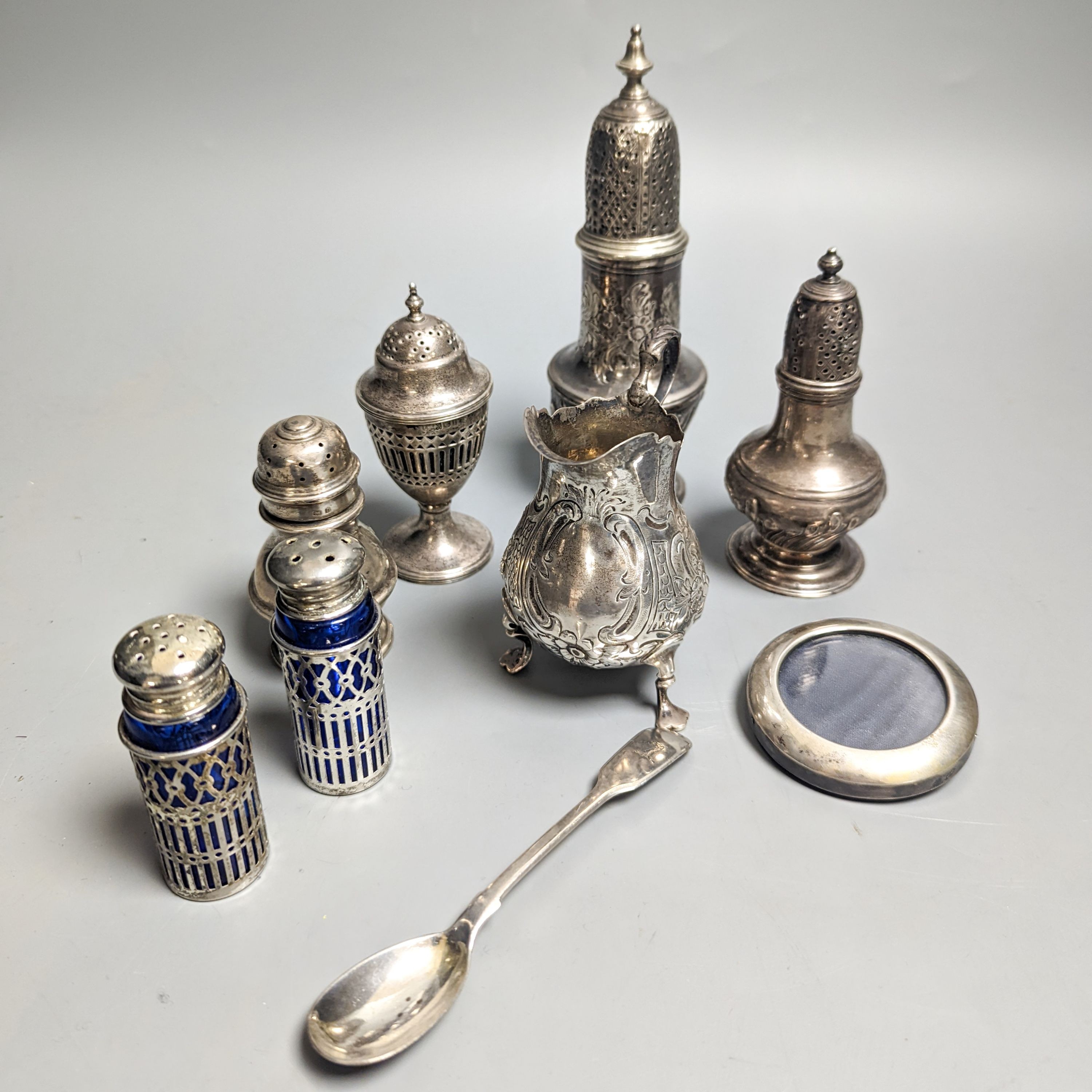 A Victorian embossed silver cream jug, London, 1883, 92mm, two 18th century silver pepperettes, one with plated cover, four later condiments (two silver), a silver spoon and small 800 standard photograph frame.